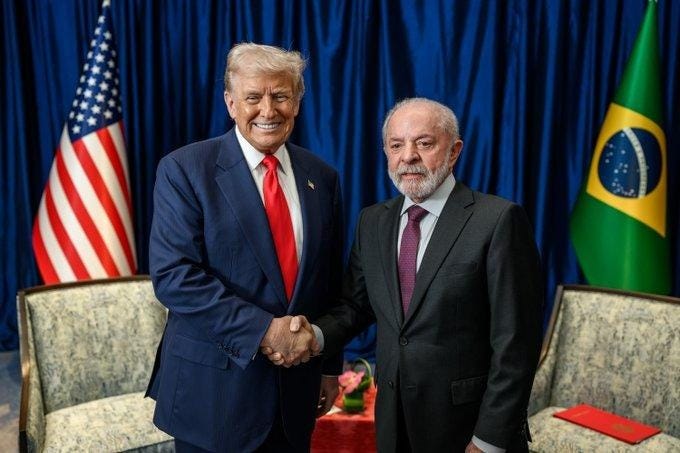 Two men in dark suits and ties stand shaking hands in a formal room setting. The man on the left has blonde hair, wears a red tie, and smiles broadly. The man on the right has gray hair, a beard, and a purple tie. Behind them is a blue curtained backdrop flanked by the American flag on the left and the Brazilian flag on the right. Two upholstered chairs are positioned nearby with a small table holding flowers and a red folder. Two men in dark suits and ties stand shaking hands in a formal room setting. The man on the left has blonde hair, wears a red tie, and smiles broadly. The man on the right has gray hair, a beard, and a purple tie. Behind them is a blue curtained backdrop flanked by the American flag on the left and the Brazilian flag on the right. Two upholstered chairs are positioned nearby with a small table holding flowers and a red folder.