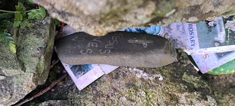 1. pebble inserted to gap between corbelled stones on the dome of the holy well that reads Jesus loves you. 2. Pebble painted in brights stripes inserted between corbelled stones that reads Smile More Often. 3. Close up of sprawling tree with large shiny green leaves with scraps of cloth, socks and all manner of oddities tied to its branches