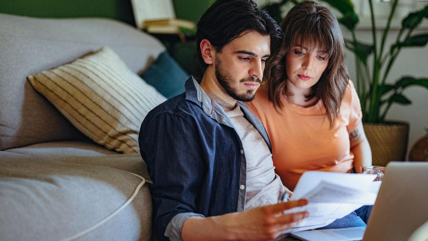Man and woman sitting on floor in front of sofa with laptop, looking at bills