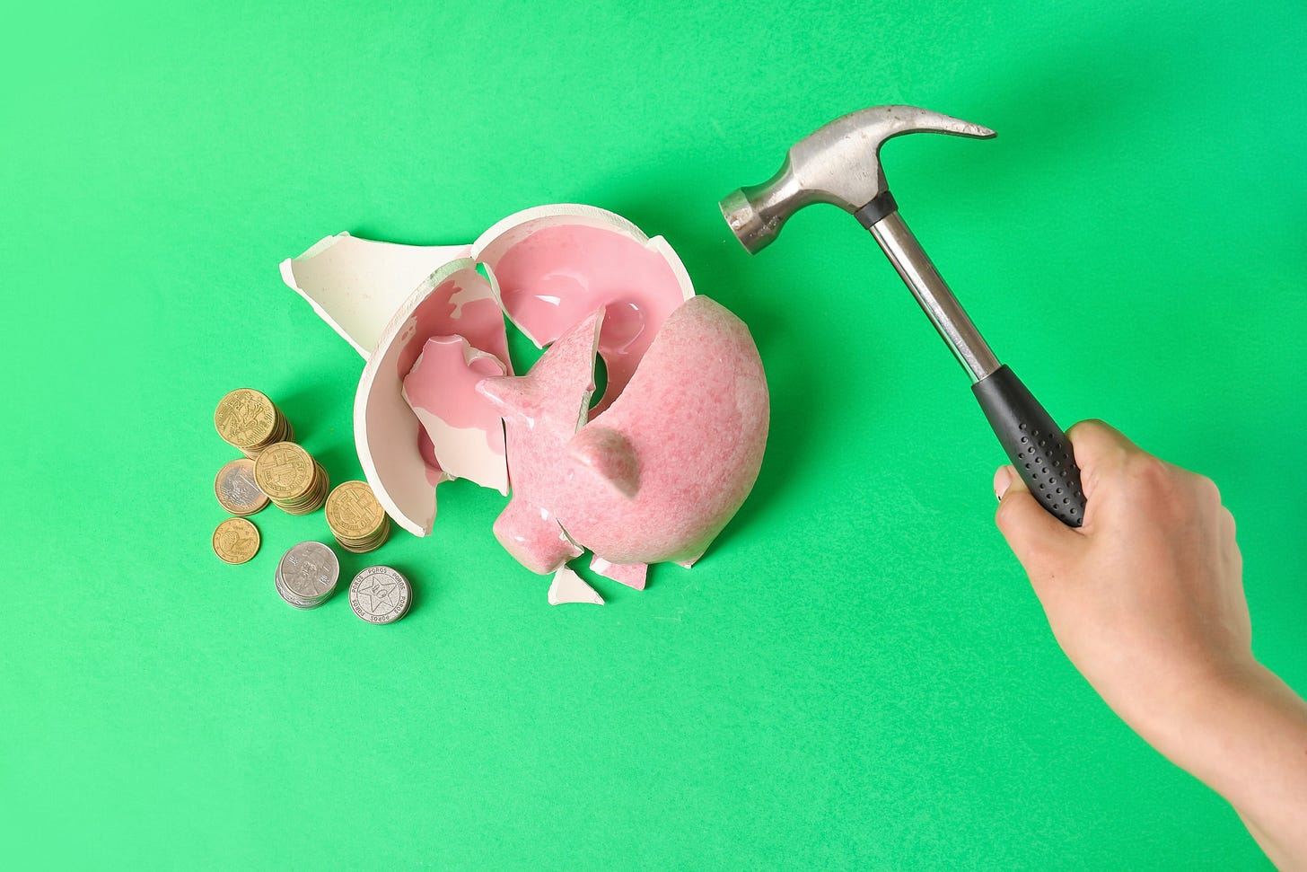 green background, a pink piggy bank has been broken open by a hammer on show with a stack of coins beside it.