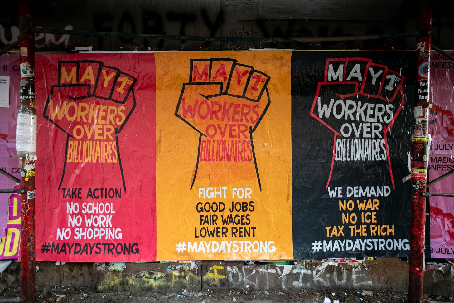 Banners hanging over a graffitied wall. From the left, a read banner with a first reads "Workers Over Billionaires." Take action. No work. No school. No shopping. #MayDayStrong. A yellow  banner with the same fist reads "fight for good jobs, fair wages, lower rent." A black banner with the same fist reads "We demand No War, No Ice, Tax the Rich." #MaydayStrong