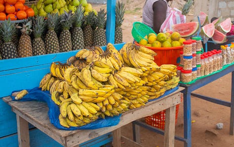 Two multicolored high rise apartment buildings, a Burger King sign near a stairwell in the mall, and a table full of fresh plantains on a roadside vendors table.