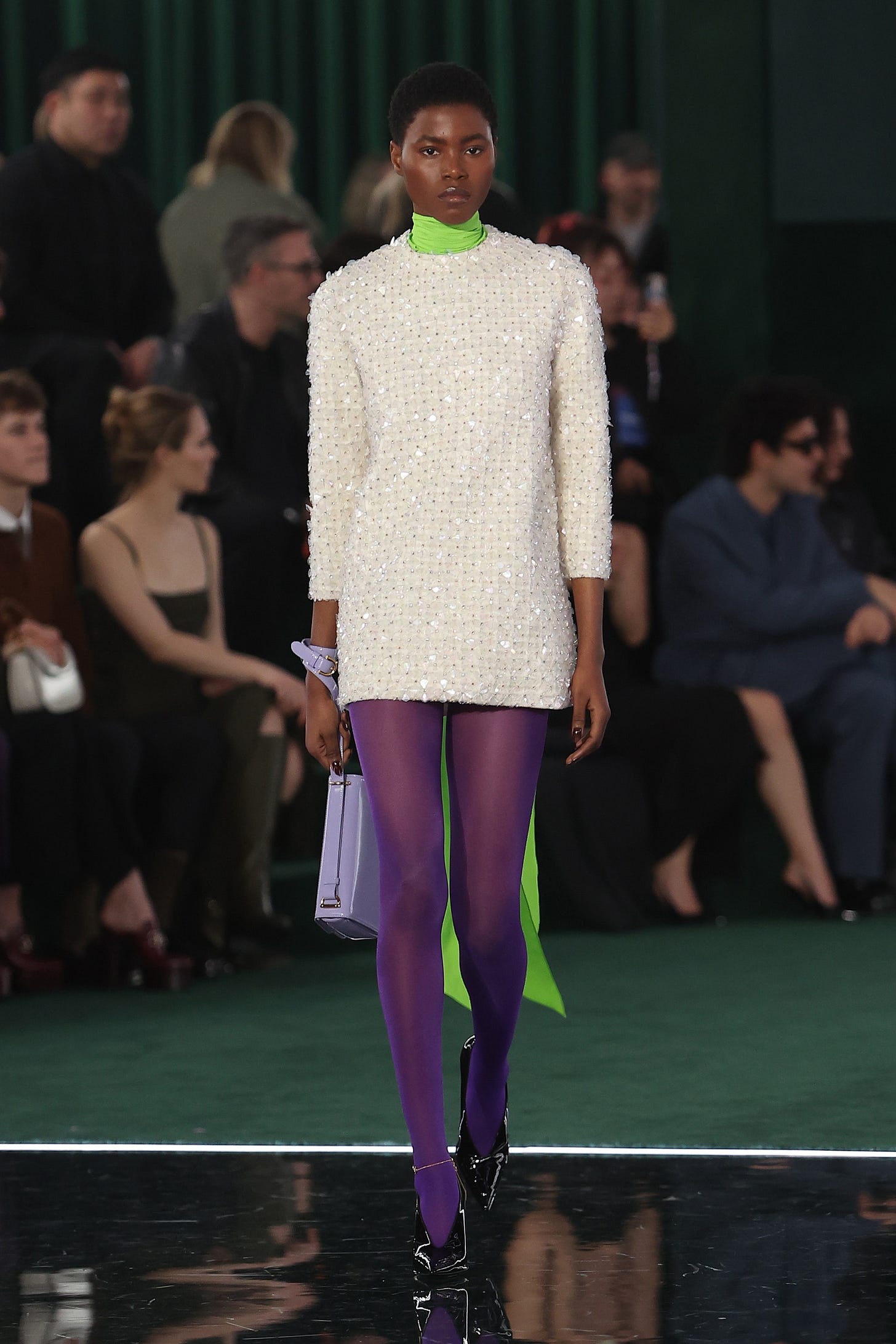 A model walks a runway wearing an embroidered off-white mini dress layered over a lime green turtleneck and purple stockings.