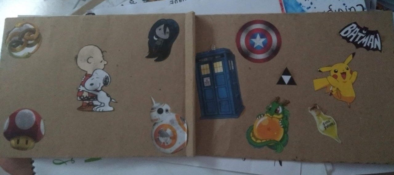 A imagem mostra capa e contracapa de um caderno coberto por papel pardo e uma coleção de adesivos e imagens de personagens e elementos de cultura pop, por exemplo: Batman, Snoopy e Charlie Brown, Capitão América, Pikachu, BB-8, Cogumelo do Mário e TARDIS.