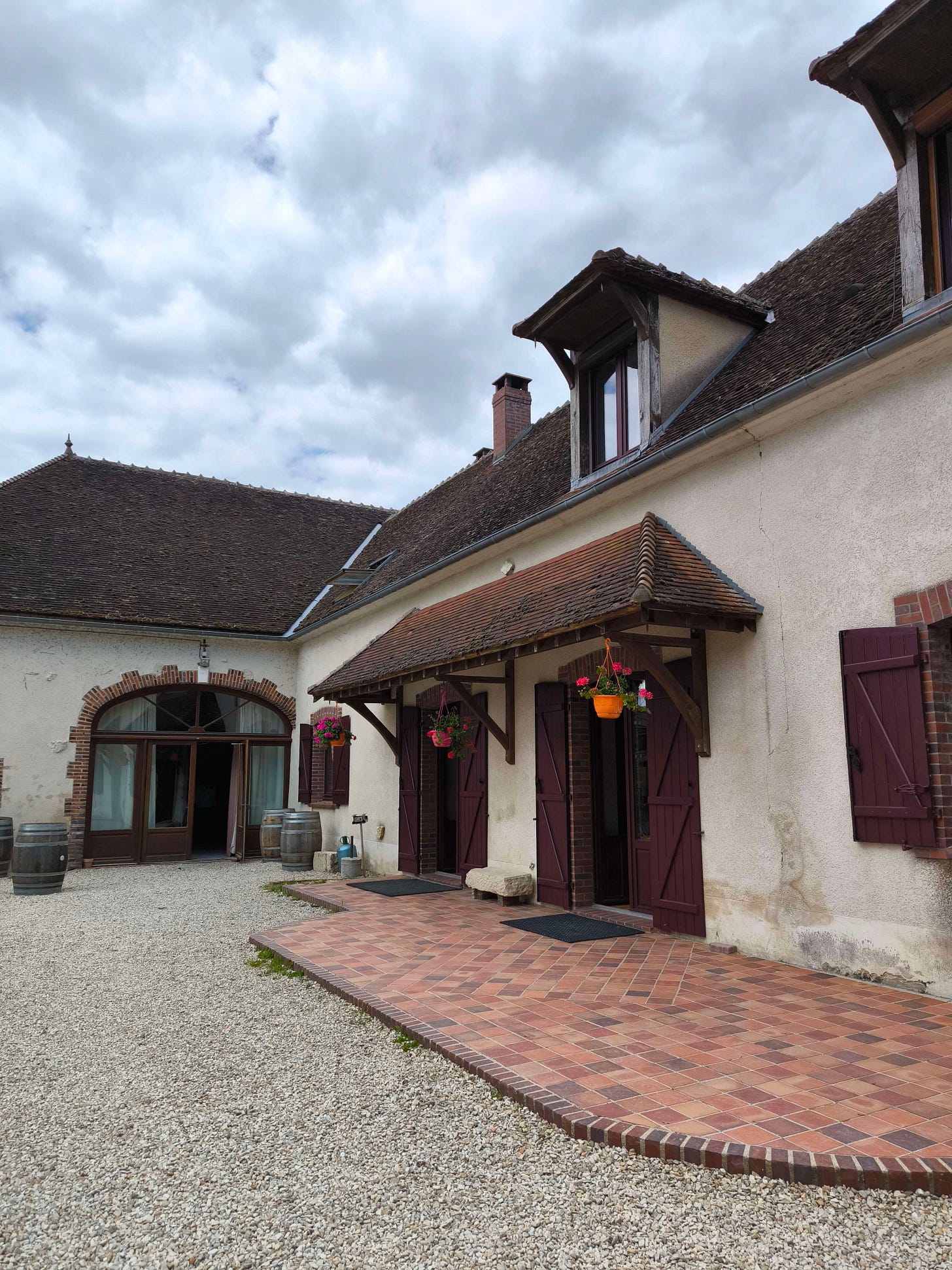 Une vieille ferme rénovée avec une terrasse pavée, des pots de fleurs roses accrochées au toit et des tonneaux.