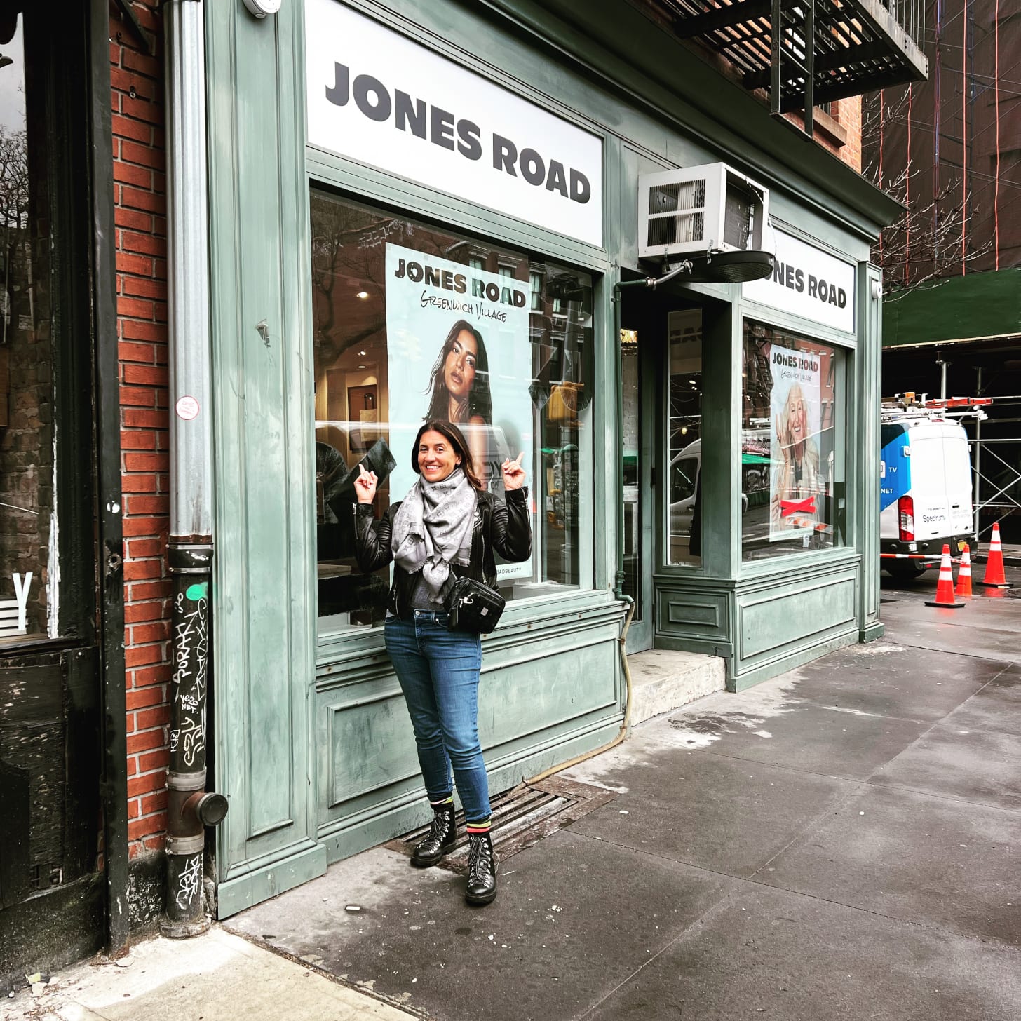 Beauty Editor Donna Francis stands smiling in front of the Jones Road Beauty storefront in Greenwich Village. She is wearing a black leather jacket, a grey patterned scarf, blue jeans, and black lace-up boots. She points enthusiastically at the shop windows, which feature large editorial posters of the brand’s "real skin" campaigns.
