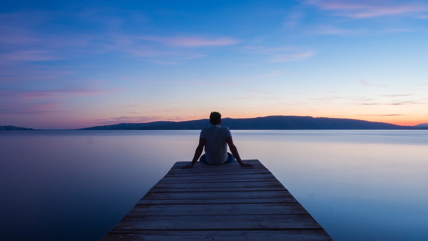 A man sits on the edge of a wooden pier overlooking a lake and mountains. The sunrise is pink on the horizon and dark blue overhead. A man sits on the edge of a wooden pier overlooking a lake and mountains. The sunrise is pink on the horizon and dark blue overhead.