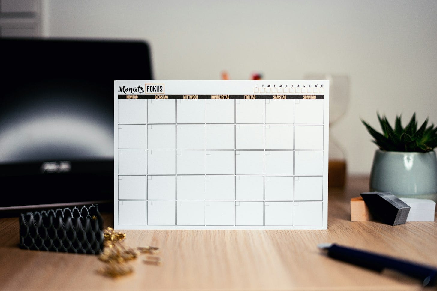 A blank monthly paper calendar stands on a wooden desk beside a potted plant, with a computer screen blurred in the background. A blank monthly paper calendar stands on a wooden desk beside a potted plant, with a computer screen blurred in the background.