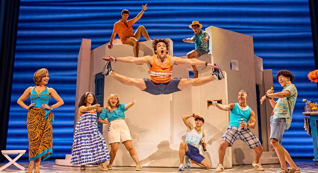A cast on stage with a man jumping in the middle and the rest posing behind him A cast on stage with a man jumping in the middle and the rest posing behind him