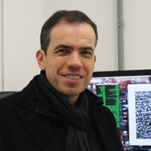 Homem de cabelos curtos sorridente usando casaco e cachecol pretos, em frente a um monitor de computador exibindo uma imagem com texturas Homem de cabelos curtos sorridente usando casaco e cachecol pretos, em frente a um monitor de computador exibindo uma imagem com texturas