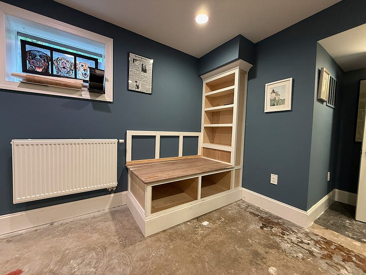 A four-photo collage showing the later stages of building a custom reading nook and bookshelf. The top-left image shows a built-in bench with a walnut top and an adjacent floor-to-ceiling bookshelf installed beneath a ceiling bulkhead. The top-right image features the partially completed bench with a cat lounging on the seat and a black dog standing in the foreground. The bottom-left image shows additional trim and paneling added to the bench back and bookshelf, with the project taking shape within the blue-walled room. The bottom-right image shows further progress on the built-in, including decorative trim, shelving, and storage cubbies beneath the bench. Together, the photos document the transformation from basic cabinetry to a finished custom reading nook.