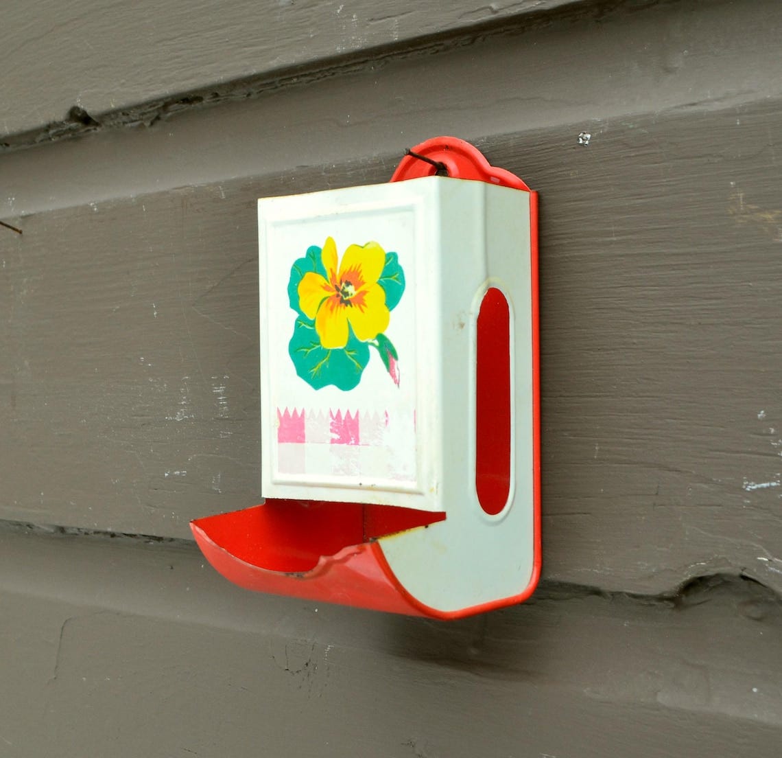 May include: Vintage metal match holder, painted white with a red trim and a red tray. The front features a yellow and green floral design. The match holder is mounted on a brown wooden surface. May include: Vintage metal match holder, painted white with a red trim and a red tray. The front features a yellow and green floral design. The match holder is mounted on a brown wooden surface.