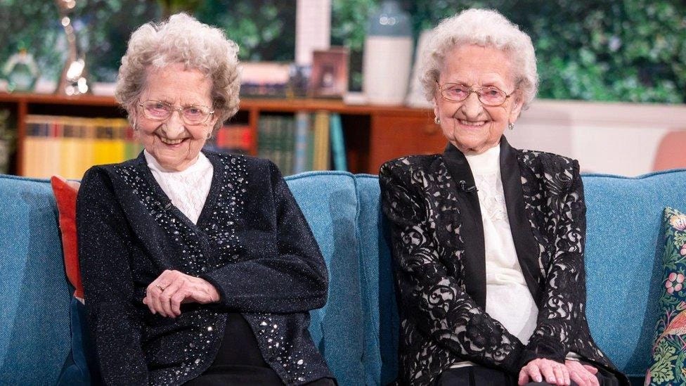 A picture of two elderly, smiling ladies in their 90's. They are twins, and look very similar. 