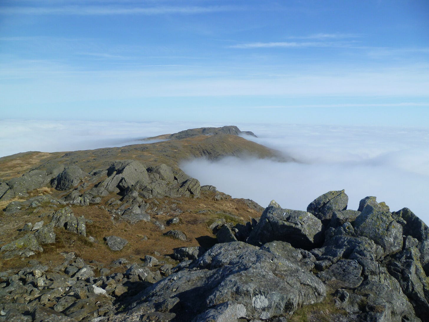 A view from a rocky mountain summit looking along a ridge that rises above a thick, white sea of clouds. Large grey boulders fill the foreground, leading to a grassy, brown mountain spine that stretches into the distance, appearing as an island above the cloud inversion under a clear blue sky.