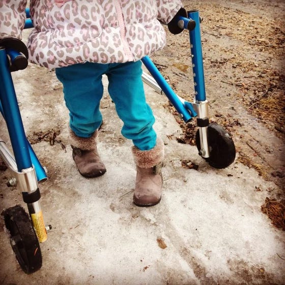 The lower half of a toddler using a walker along a dirt-road packed with some ice and snow.