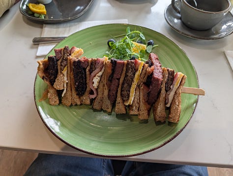 A Japanese Strawberry Shortcake triple layer cake with cream frosting and strawberry decoration, a club sandwich full of cooked breakfast items, and an adjustable angle art desk
