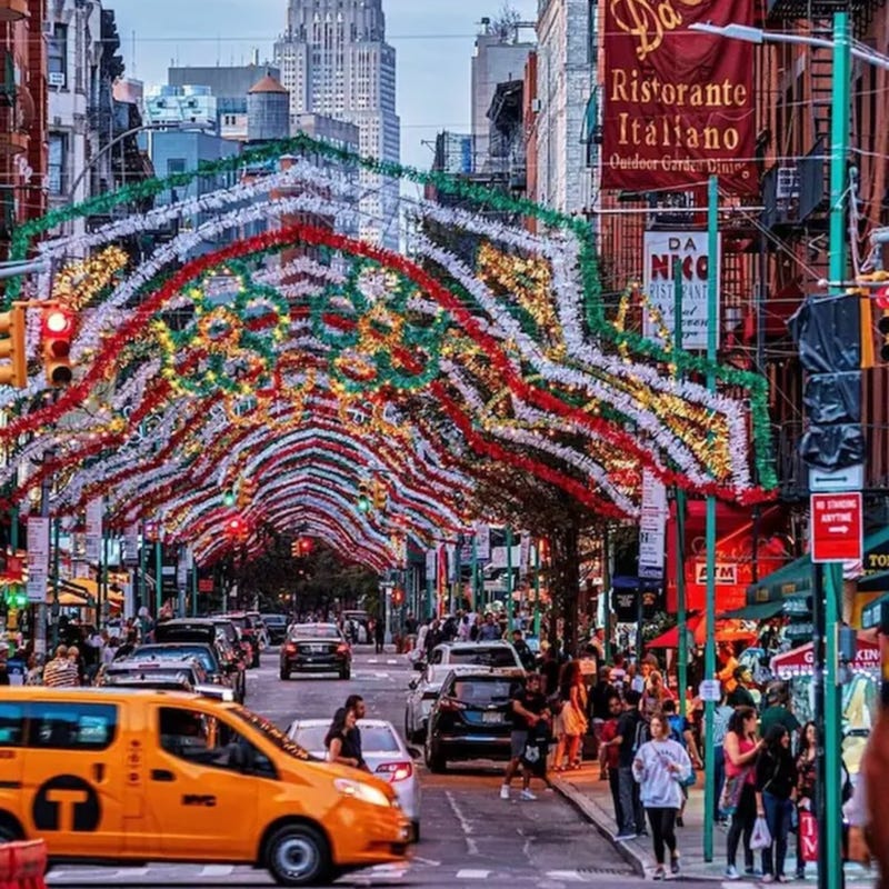 alt="Feast of San Gennaro celebration in Little Italy NYC with festive lights, crowds, Italian eateries, and city traffic"