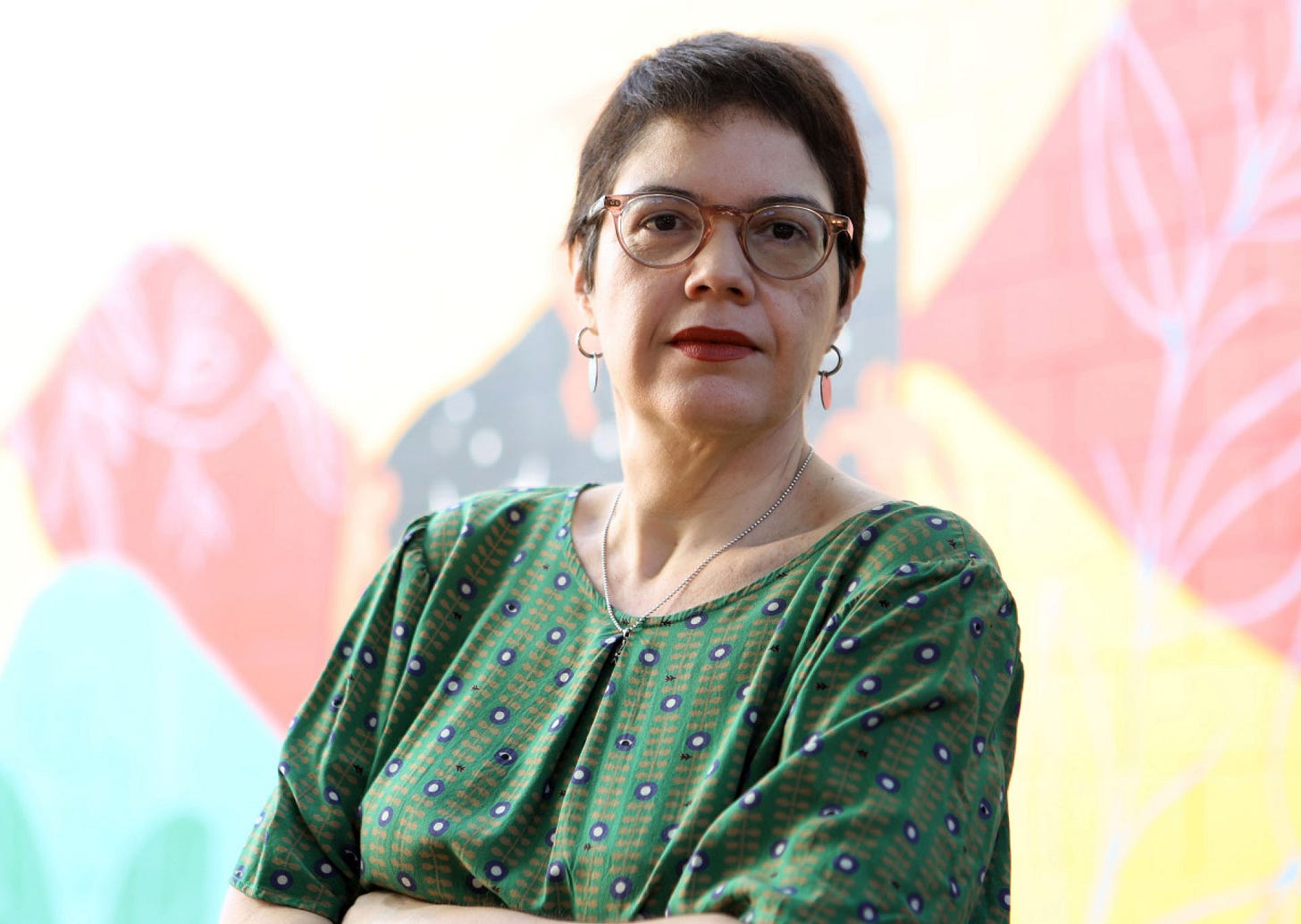 Pessoa usando óculos e vestindo blusa verde estampada, posicionada em frente a um fundo com elementos decorativos em tons de rosa, amarelo e verde.
