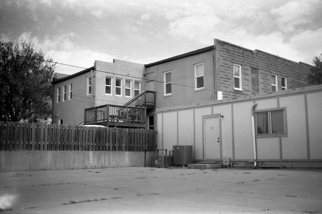 A black and white photograph of the back of a building, with stairways and a wooden fence