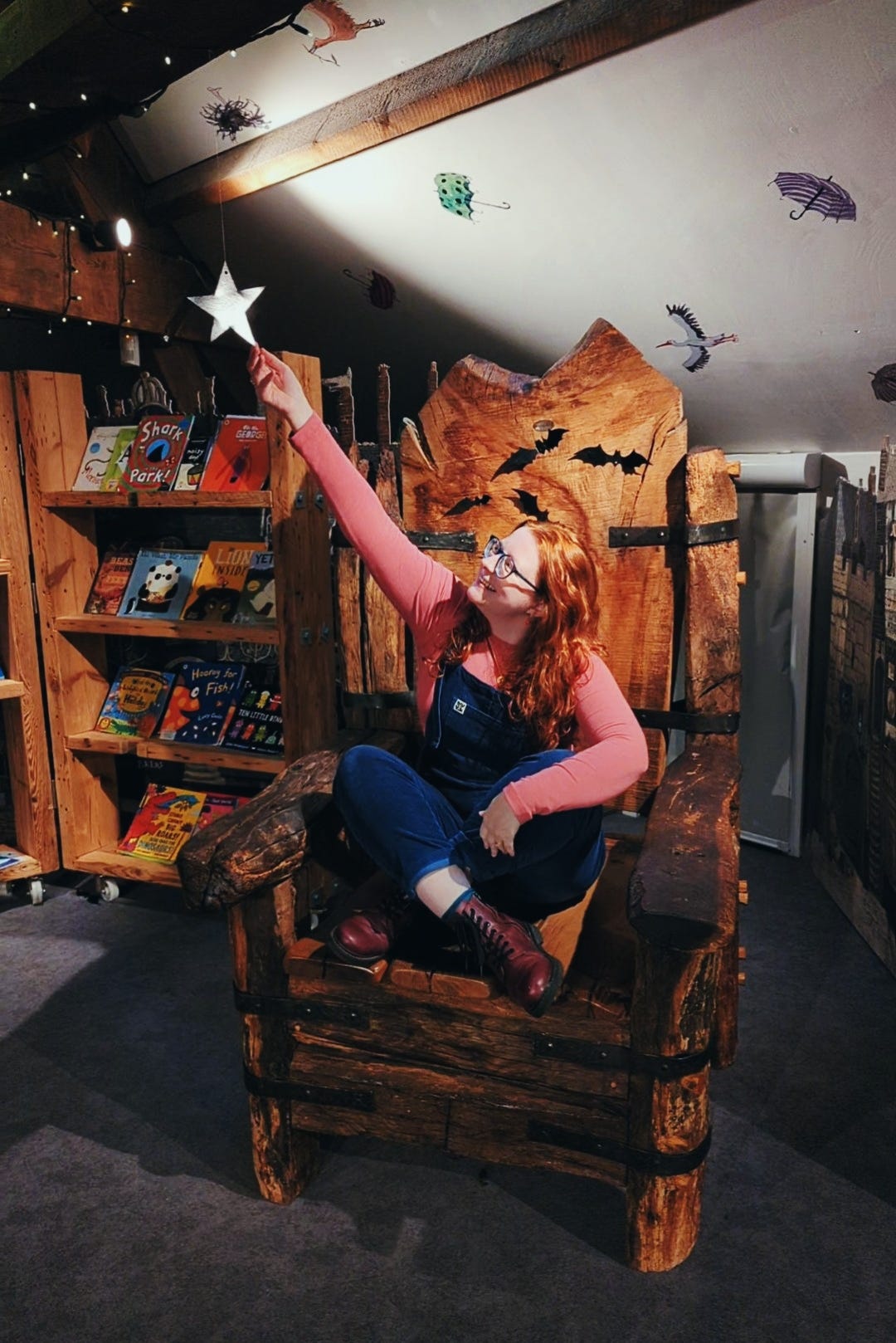 A white woman with long red hair and blue glasses sits cross-legged on an over-sized oak throne. She is smiling and reaching her hand up to touch a shining star, hanging from the fairy-lights adorning the ceiling beams above her. A collection of children's books sit on shelves next to her. She is wearing a pink, long-sleeve top, navy, cord dungarees and purple boots. 