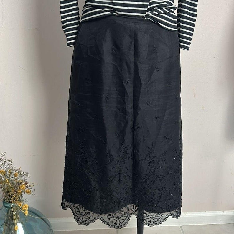 May include: Black midi skirt with embroidered floral details and a scalloped lace hem. The skirt is made of a lightweight fabric and has a subtle sheen. It is paired with a black and white striped long-sleeve shirt. The skirt is displayed on a mannequin.