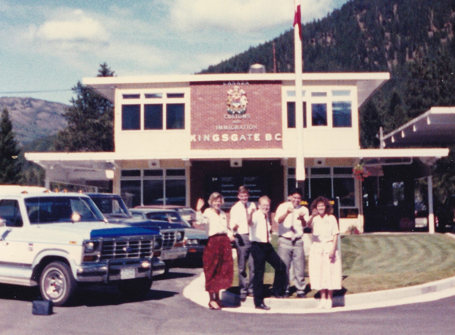 Five young LDS missionaries (two women and three men) stand at the grassy edge of a parking lot in front of the Canadian Customs and Immigration station in Kingsgate, British Columbia. Behind them rises a wooded slope, and beyond that fluffy clouds ride a light blue sky. Five young LDS missionaries (two women and three men) stand at the grassy edge of a parking lot in front of the Canadian Customs and Immigration station in Kingsgate, British Columbia. Behind them rises a wooded slope, and beyond that fluffy clouds ride a light blue sky.