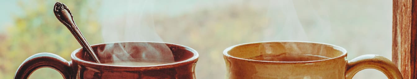 Two steaming mugs of hot beverages sit on a windowsill with a blurred autumn background.