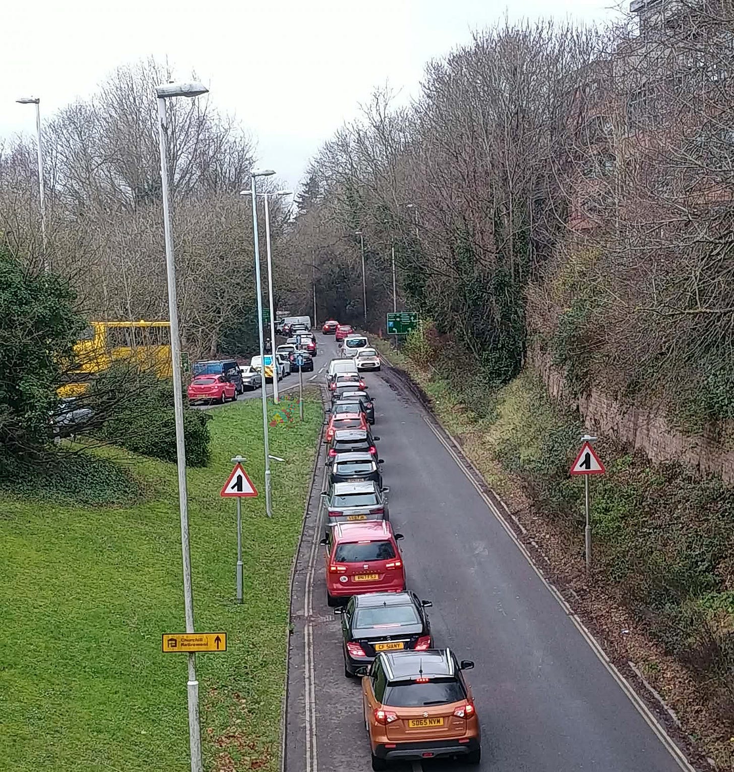 Traffic jam on the A22 merging with traffic from East Grinstead town centre.  lanes are divided by winter trees.  Traffic is bumper to bumper near the merge sign and sharp bend warning.  Streetlights and grassy verges line the road as vehicles wait in heavy congestion.