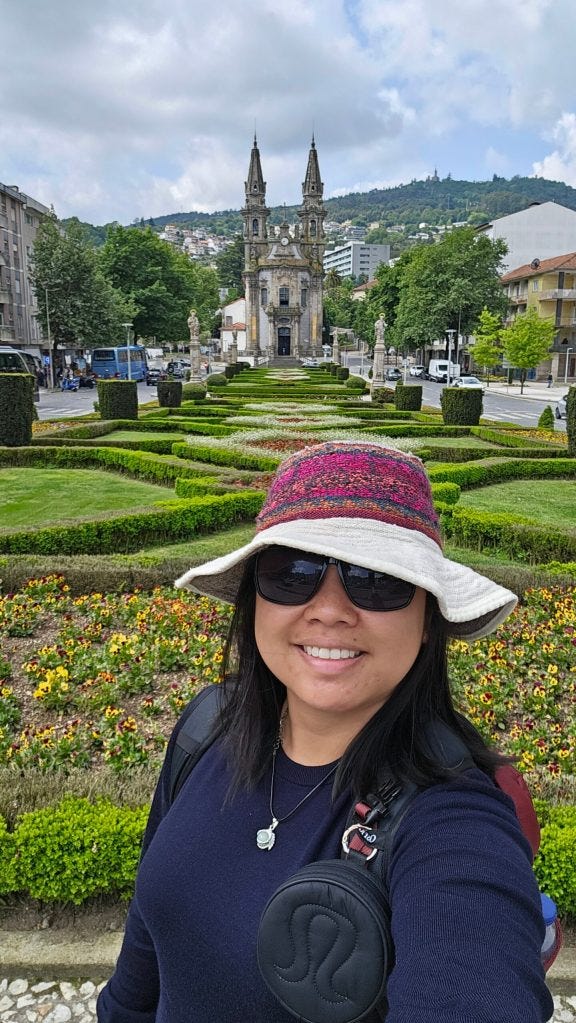 A person taking a selfie in a park with manicured gardens and a church in the background, dressed casually with sunglasses and a wide-brimmed hat. A person taking a selfie in a park with manicured gardens and a church in the background, dressed casually with sunglasses and a wide-brimmed hat.