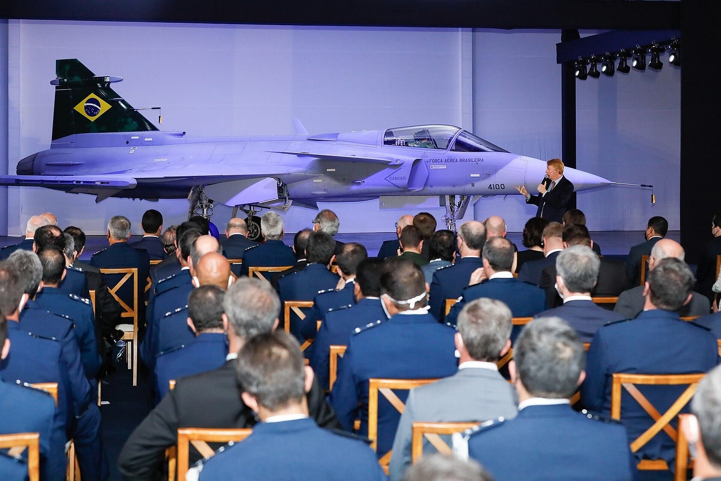 A man speaks on a stage beside a grey Gripen fighter lit in blue, facing rows of seated officers in blue uniforms.