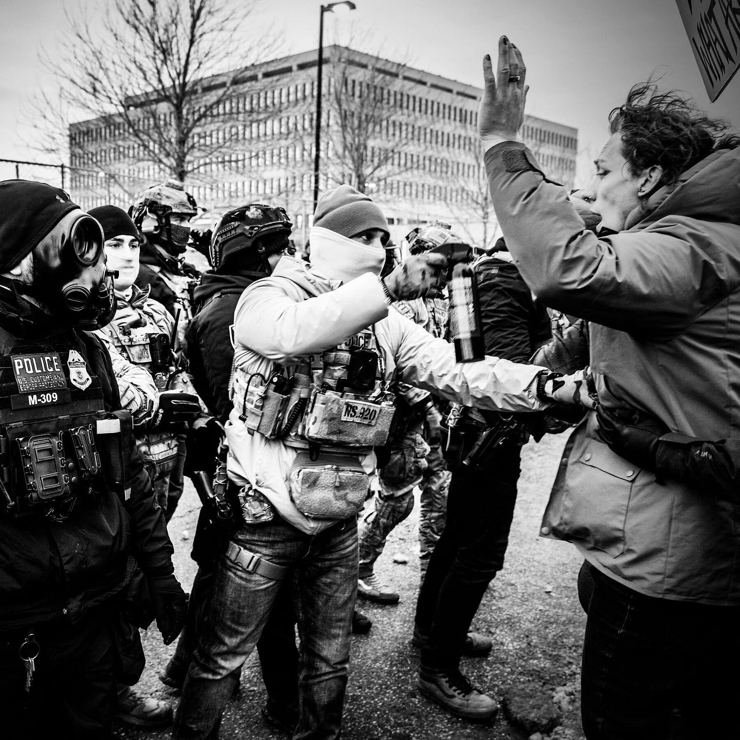 Several officers in uniform, some wearing masks, facing a demonstrator whose hands are in the air. Several officers in uniform, some wearing masks, facing a demonstrator whose hands are in the air.