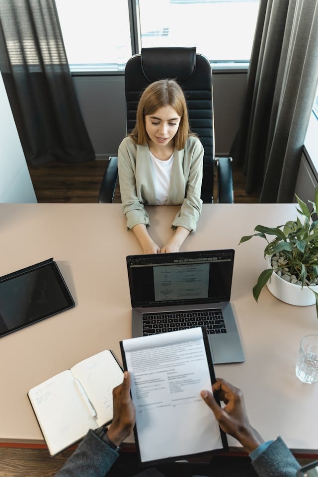 A woman speaking at a table with her hands on a table, while a person is holding a notepad with their resume on it (in addition to a laptop) A woman speaking at a table with her hands on a table, while a person is holding a notepad with their resume on it (in addition to a laptop)