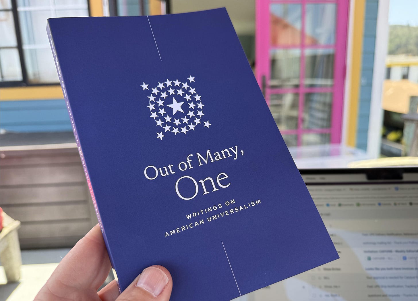 A navy blue book titled "Out of Many, One" with a logo of white stars arranged in a circular pattern. The subtitle reads "Writings on American Universalism". The book is being held by a hand, with a blurry background of windows and interior space visible. The book's design is minimalist, with white text on the deep blue background, creating a clean and scholarly appearance.