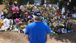 An individual with a blue shirt and black stands in front of a memorial with flowers at a roadside.