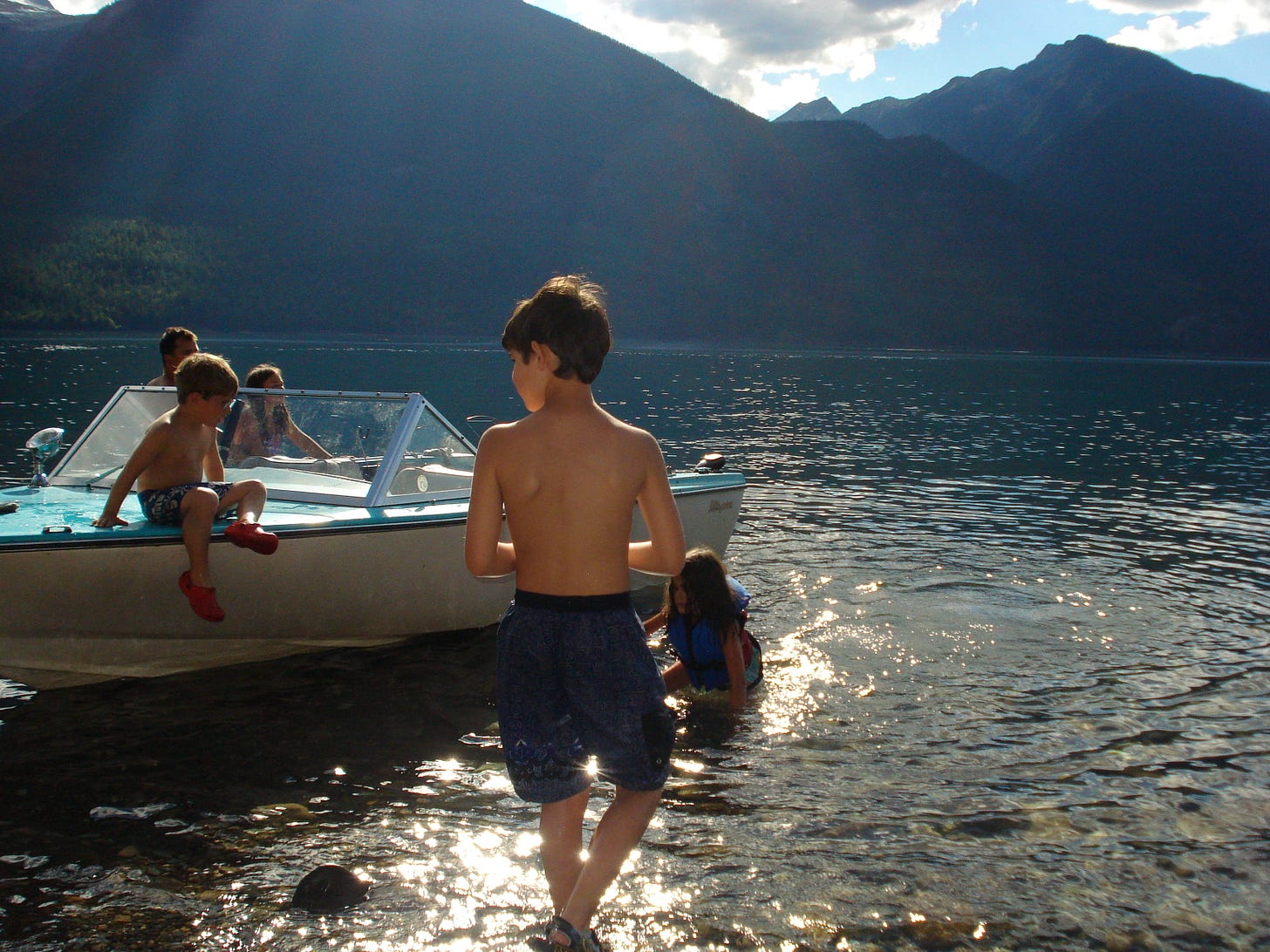 A boy stands with his back to the camera, facing a lake and mountains. young children climb on the boat.