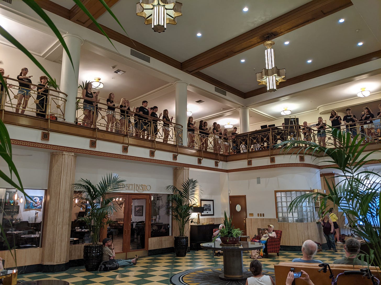 A group of 30 or so teenage singers singing from the second story balcony inside the lobby of an ornate historic building. A group of 30 or so teenage singers singing from the second story balcony inside the lobby of an ornate historic building.