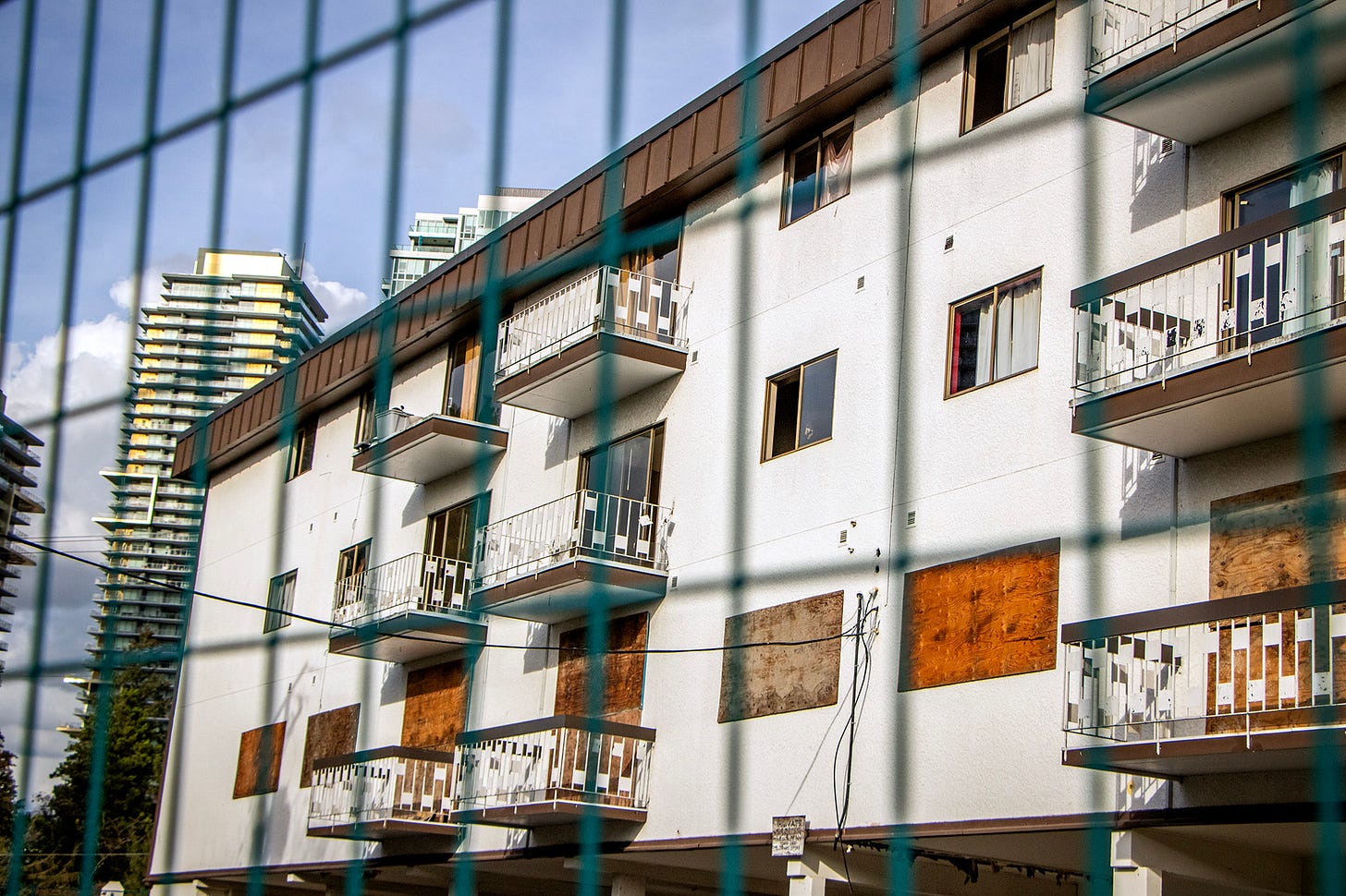 a small walk-up apartment building is seen through out-of-focus green metal fencing. the white walls are punctuated with windows and balcony doors that are boarded up