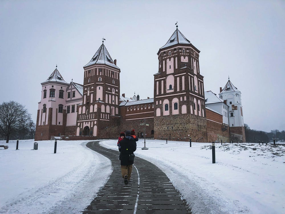 Mir Castle, Belarus. A man carrying two small children walks a cleared path through snow towards a castle of many wide turrets.