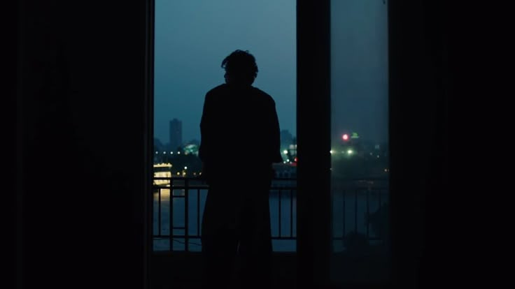 This may contain: a man standing in front of a window looking out at the city lights and water This may contain: a man standing in front of a window looking out at the city lights and water