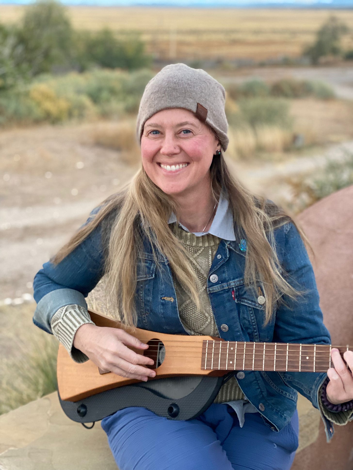a white woman wearing a jeak jacket, sweater, playing a paddle guitar in the New Mexico desert