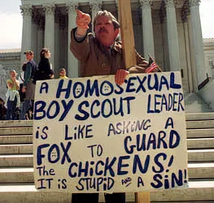 A man on the steps of the U.S. Supreme Court holding a handwritten sign that reads "A homosexual Boy Scout leader is like asking a fox to guard the chickens! It is stupid and a sin!" He is pointing at the camera and holding a small American flag.
