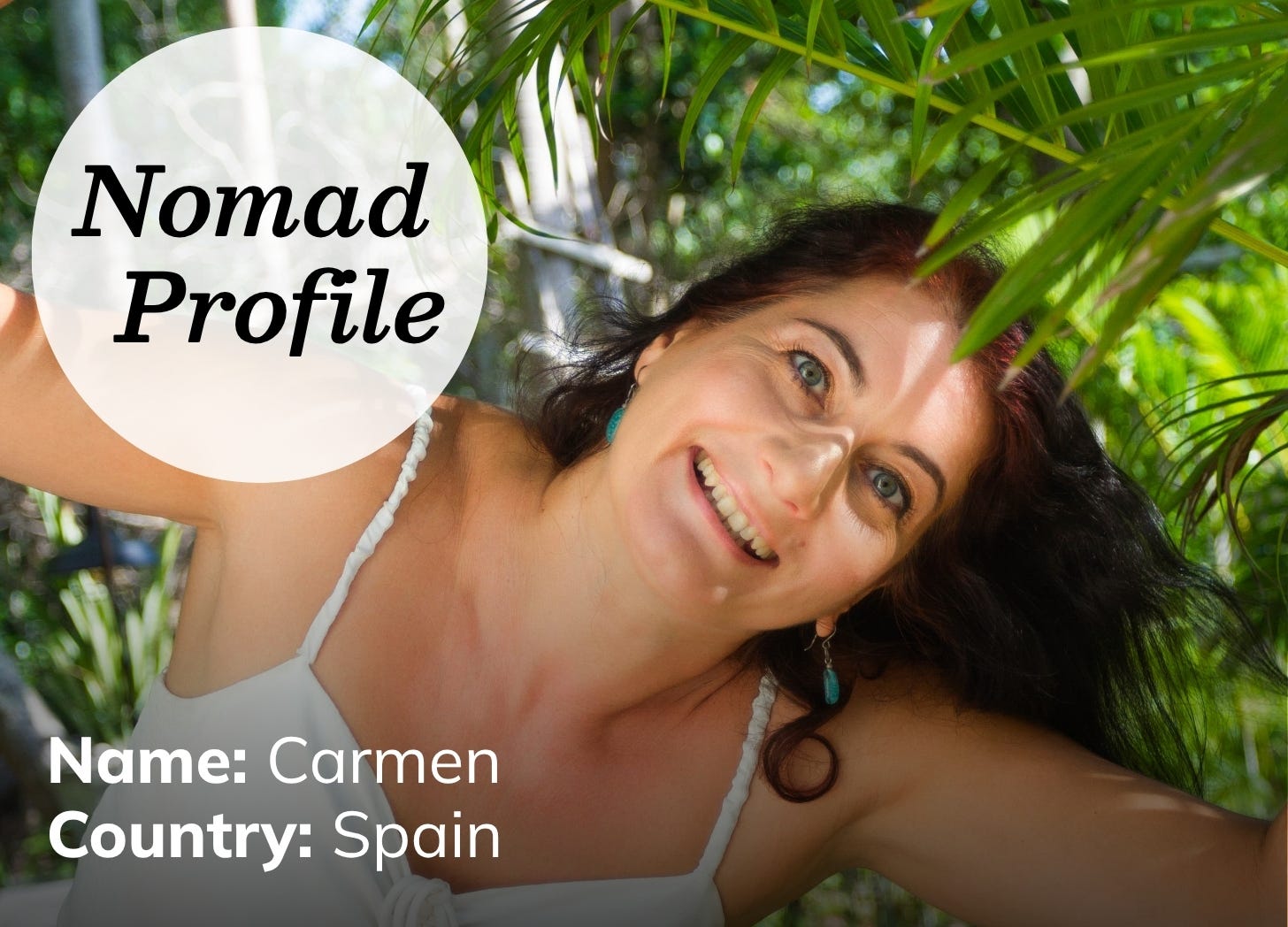 White woman with long dark hair and light eyes is smiling and wearing a white tank top. She is surround by lush green foliage. 