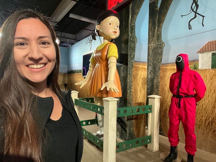 Kirsten Acuna smiles in front of the Squid Game Red Light Green Light girl and a model of a game facilitator.