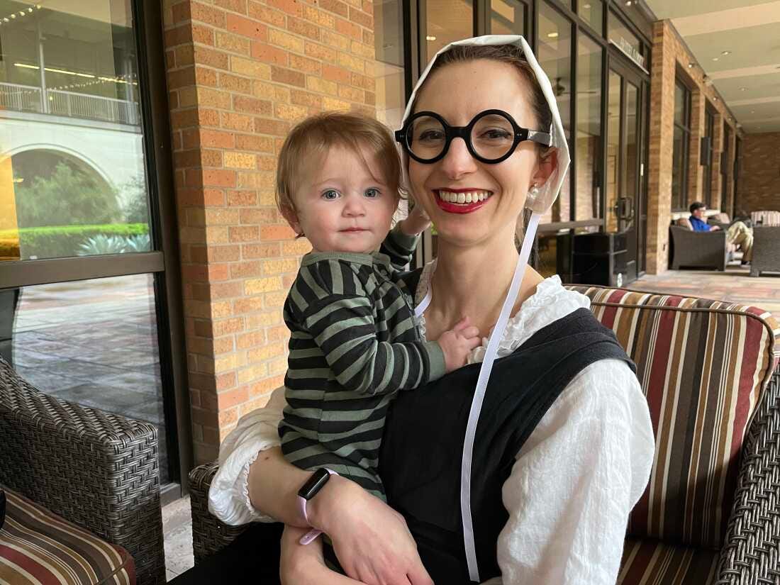 Simone Collins holds her 1-year-old daughter. Collins is wearing round, black-framed glasses and a white bonnet.