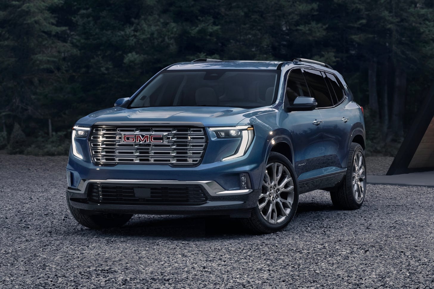 A blue 2024 GMC Acadia Denali is parked on gravel near a forest.