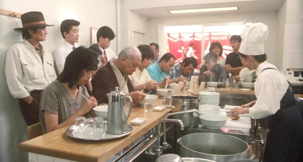 A crowded ramen shop with patrons sitting at a counter, eagerly eating noodles. A chef in white serves them.