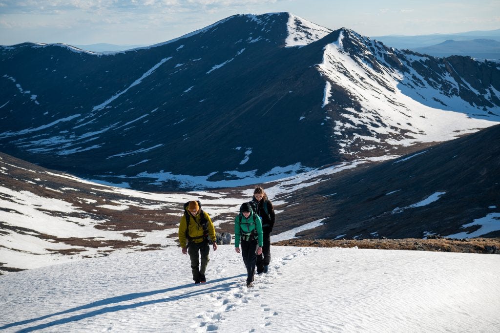 Three hikers climb a broad snow slope toward the camera in low light, a large snow-streaked bowl and dark peaks rising behind them.