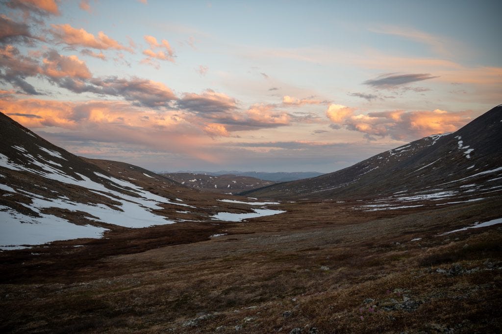 A broad U-shaped valley of brown tundra and snow patches under a twilight sky, clouds lit peach and pink by a low sun, faint ranges at the far end.