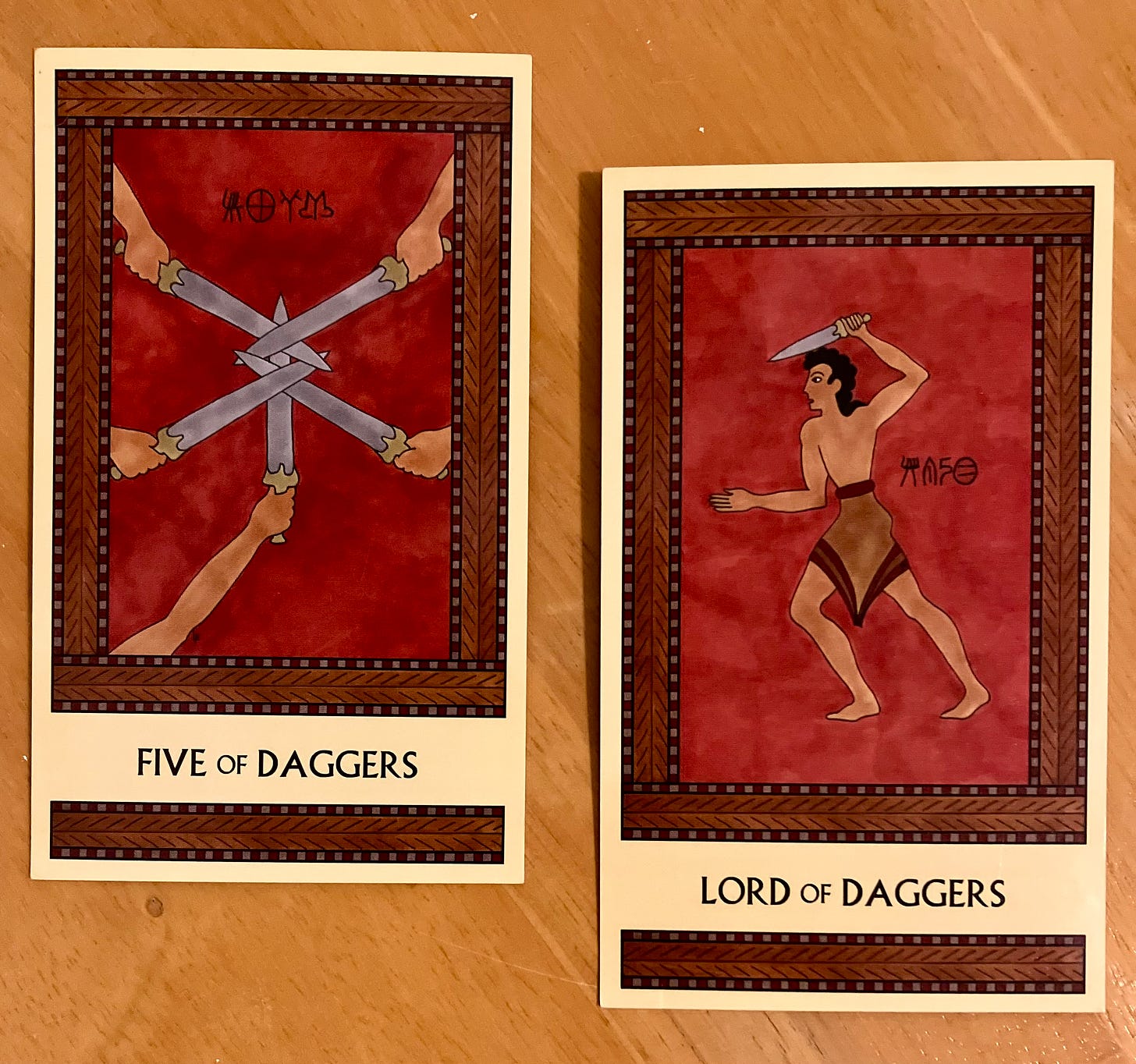Two Minoan Tarot cards side by side on a light wood surface. Both cards are in shades of dark red and brown. The Five of Daggers depicts five hands, each one wielding a long dagger. The points of the daggers are interlocked in the center of the image so none of them can be used. The Lord of Daggers shows a Minoan man wearing a short kilt, facing left, with his left arm raised above his head. He is gripping a dagger in that hand.