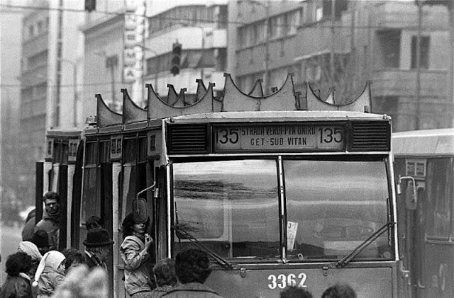 A black-and-white photograph of a crowded public bus in an urban setting. The bus displays route number "135" with the destination signs reading "Strada Verde Piața Unirii" and "CET-Sud Vitan." The bus's exterior is weathered, and passengers are seen boarding, some peering out from the open doors. In the foreground, a group of people, wearing winter clothing, waits to get on, while the cityscape in the background is hazy, hinting at a cold and foggy day. The image captures a sense of daily life and community amidst a gritty, urban atmosphere.